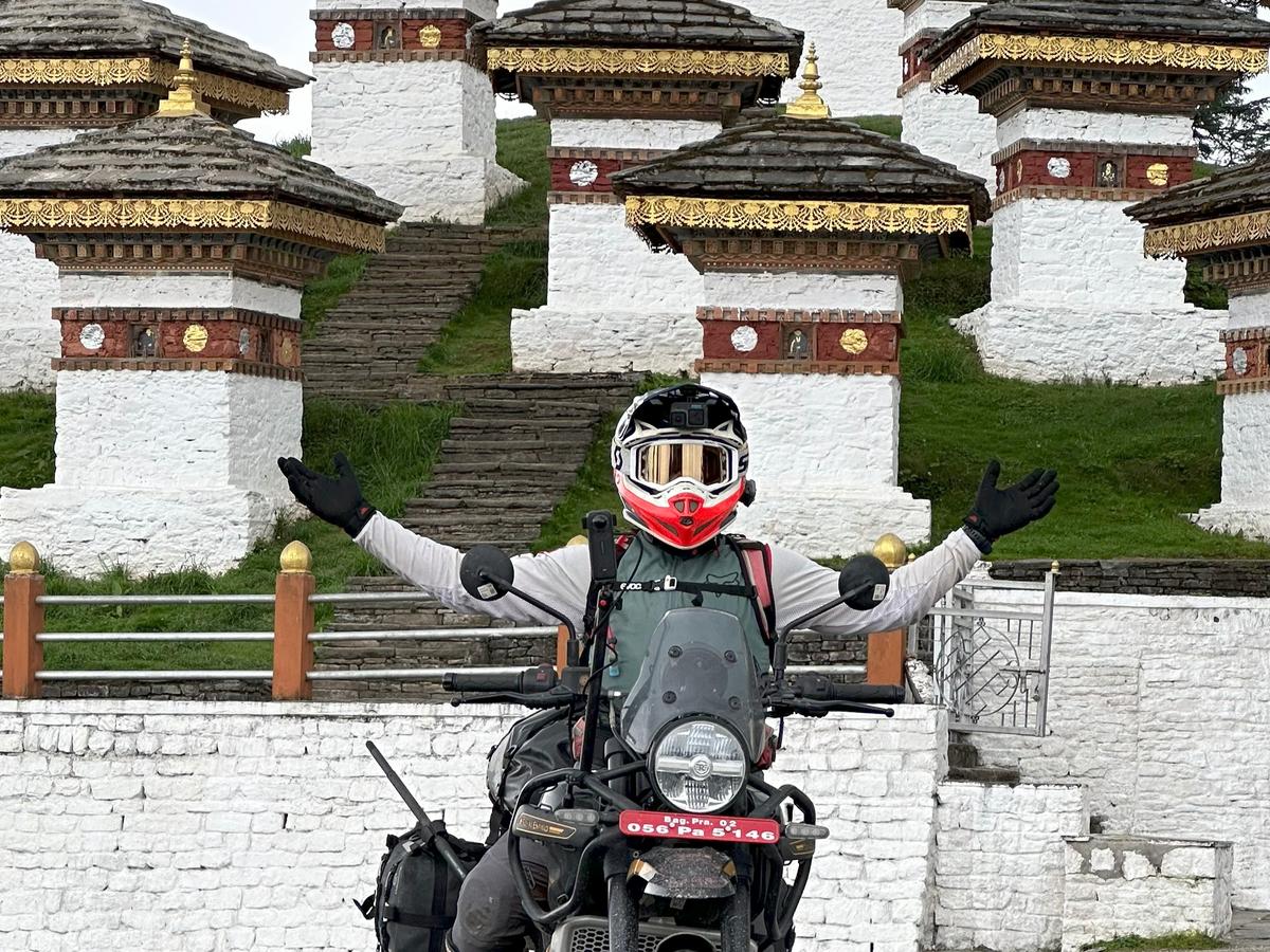 A person on a motorcycle in front of a series of white and red stupa-like structures in the mountains of Bhutan