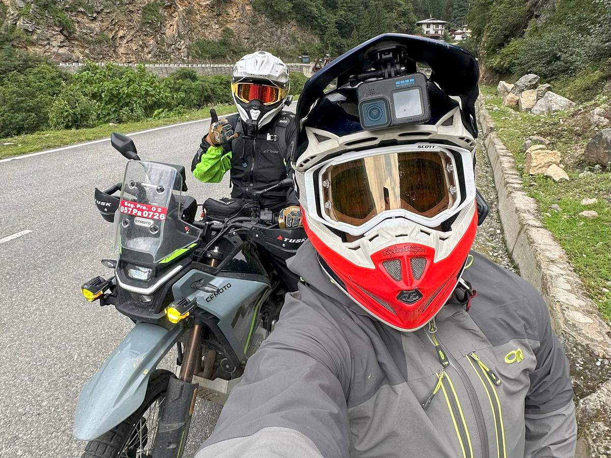 Two motorcyclists on a mountainous road, wearing helmets and riding gear, with a camera on one of the helmets