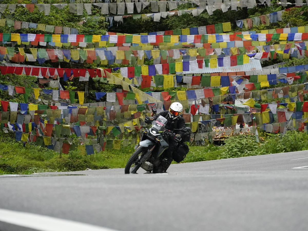 A person on a motorcycle navigating a winding road through a mountainous region, surrounded by prayer flags and trees