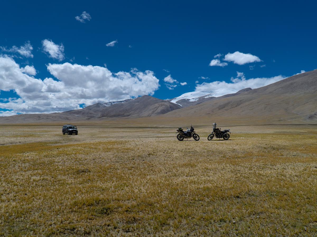 A group of motorcyclists and a 4x4 vehicle are seen in the distance, with snow-capped mountains in the background, set against a blue sky with white clouds