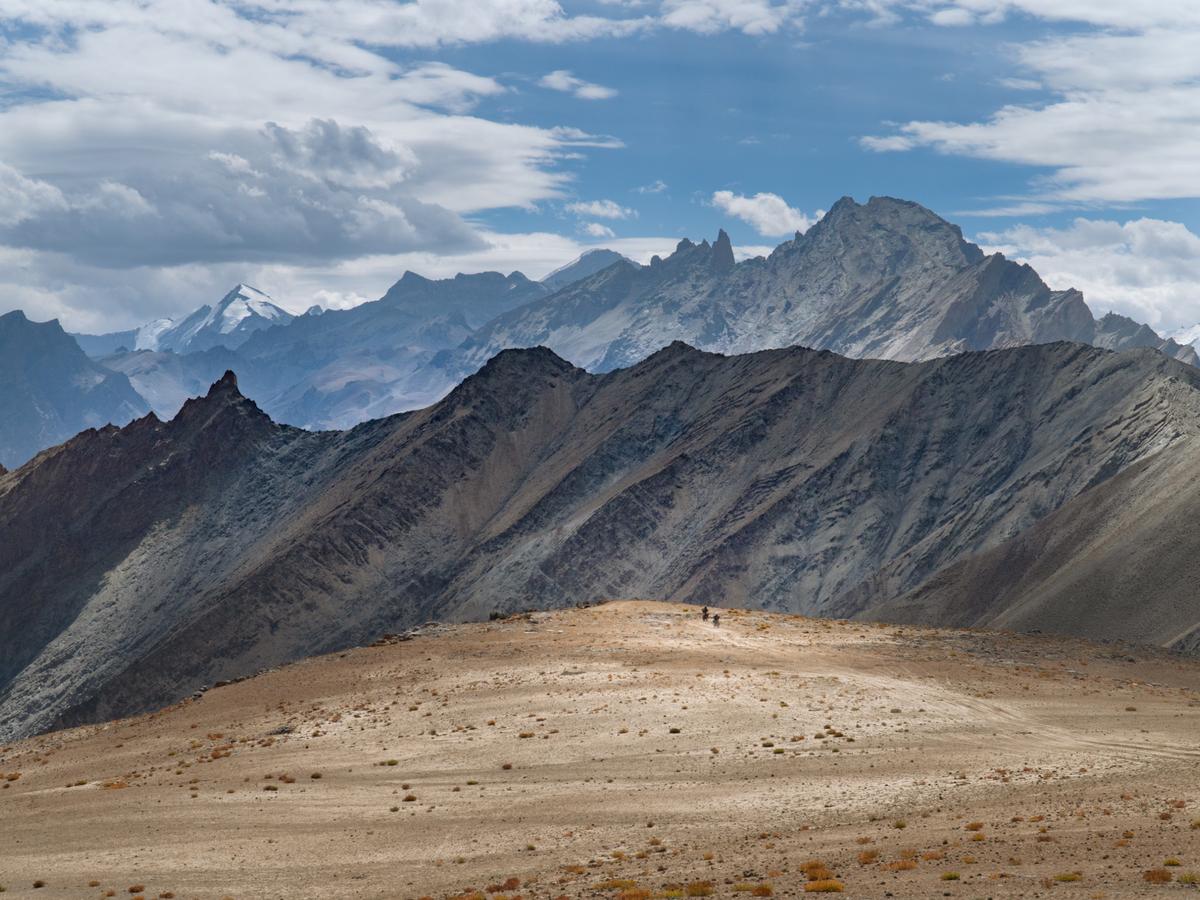 A vast, arid mountain range with two adventure moto riders in the distance, set against a blue sky with white clouds.