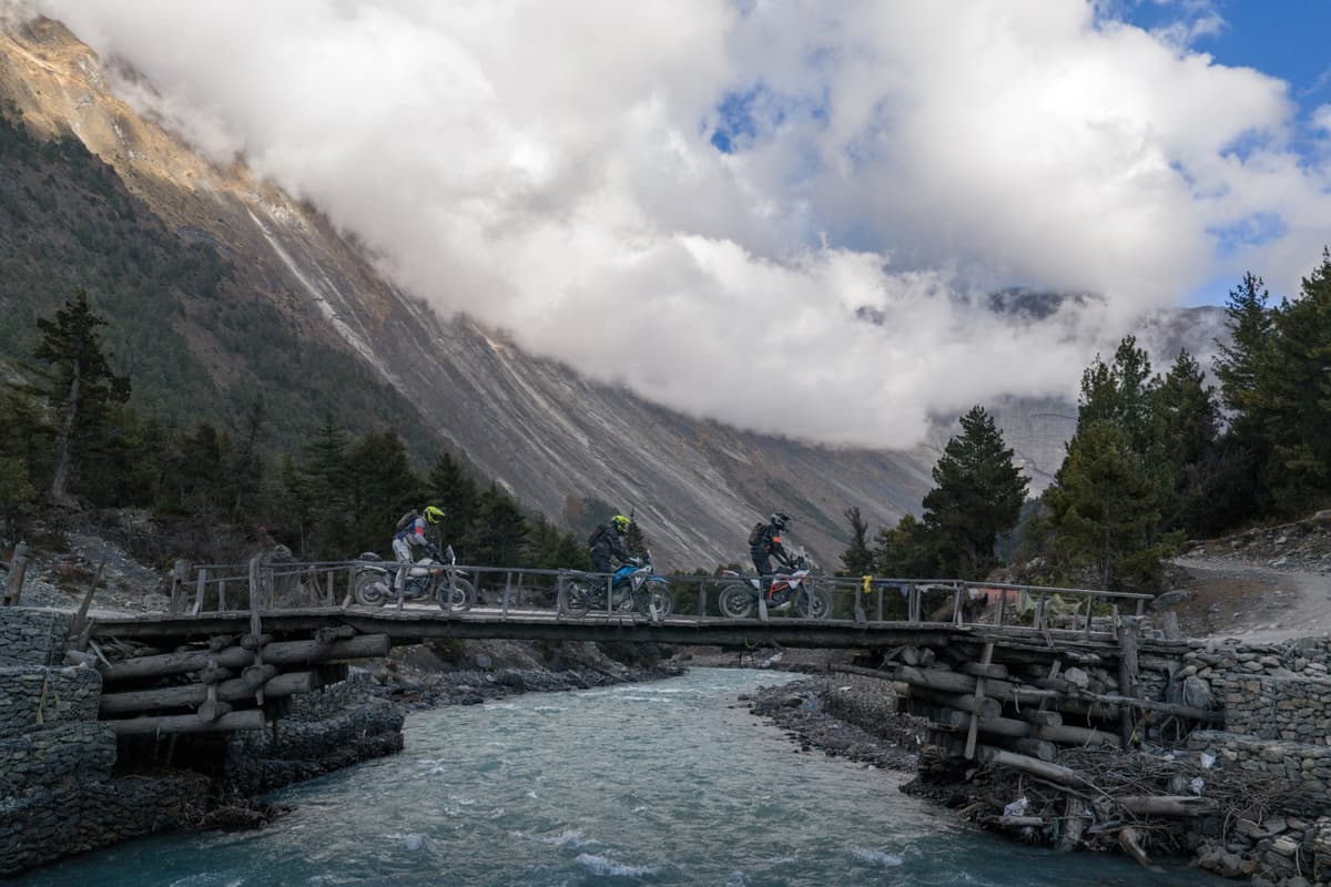 Motorcyclists riding across a wooden bridge over a mountain river during our Chris Birch Signature tour in Nepal, with pine forest and dramatic mountains in the background.