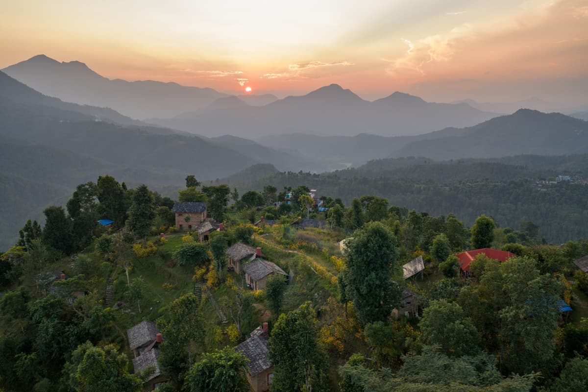 Aerial view of a hillside village surrounded by lush greenery and terraced houses at sunset with misty mountains in the background during our Nepal motorcycle tour.