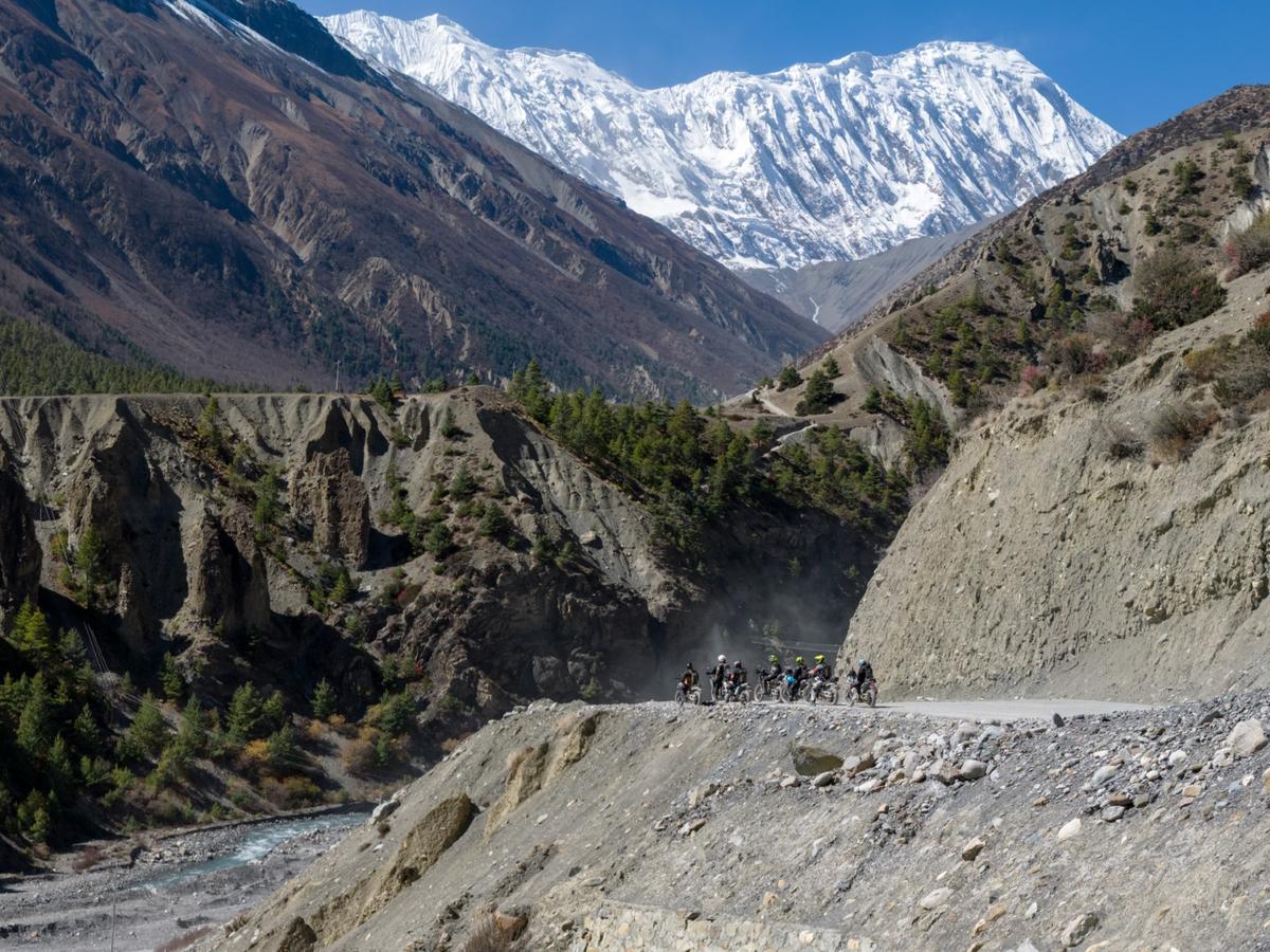 Group of motorcyclists riding along a cliffside dirt road with snow-capped mountains towering in the background during our Nepal motorcycle tour.
