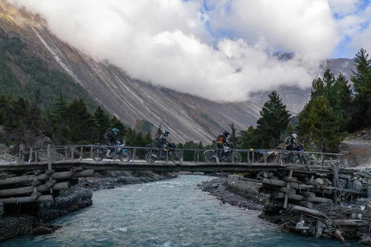 Motorcyclists riding across a wooden bridge over a turquoise river in a mountainous valley during our Nepal motorcycle tour.