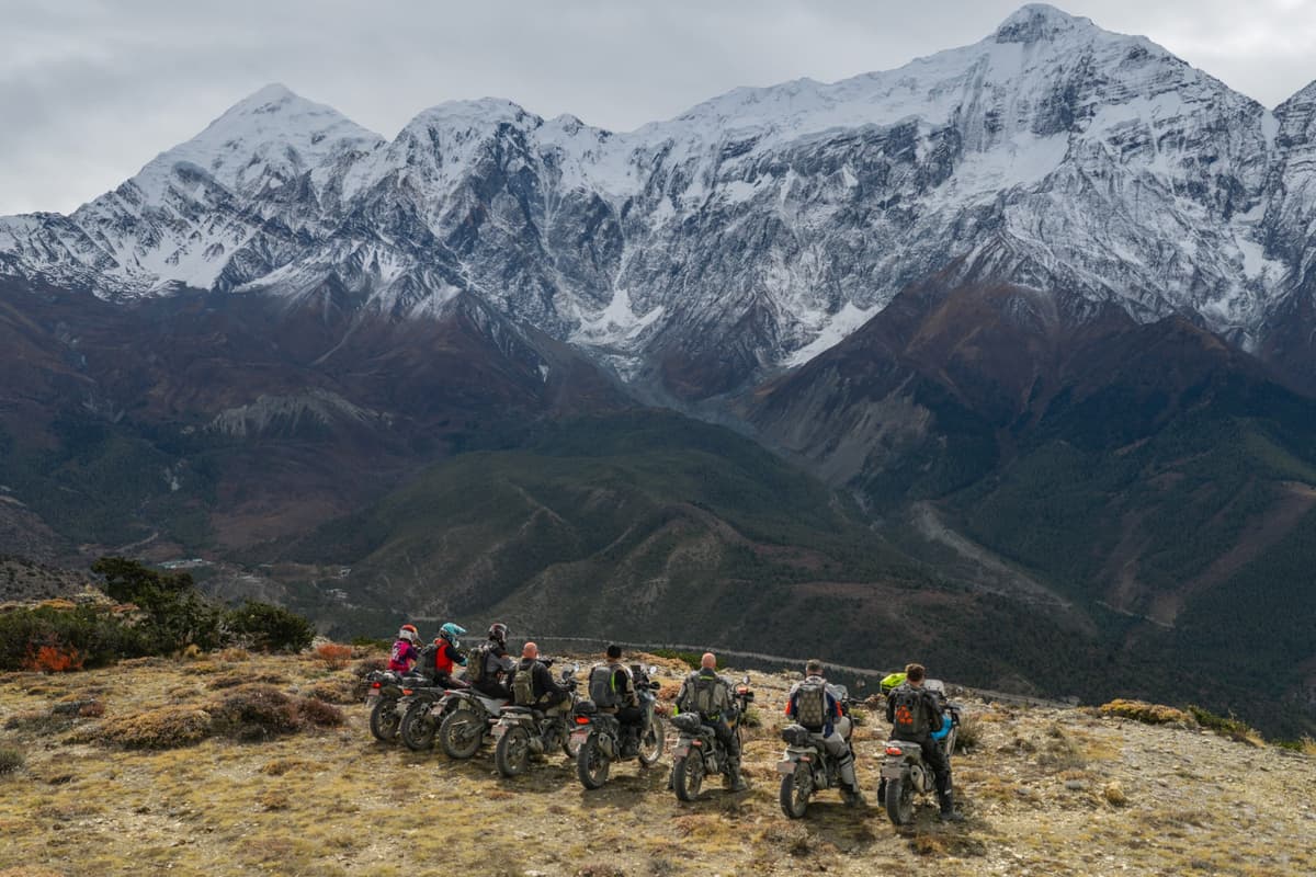 Group of motorcyclists lined up on a grassy ridge overlooking a dramatic snow-capped mountain range during our Nepal motorcycle tour.