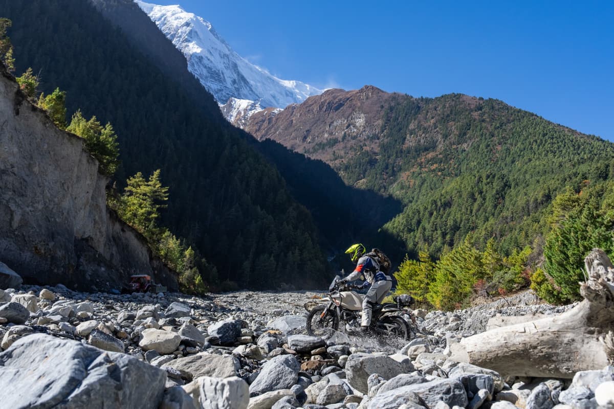 Motorcyclist riding across a rocky riverbed with large boulders and logs, with forested mountains and snow-capped peaks in the background during our Nepal motorcycle tour.