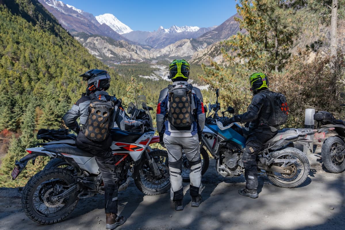 Three motorcyclists standing beside their adventure motorcycles overlooking a lush valley with snow-capped mountains during our Nepal motorcycle tour.