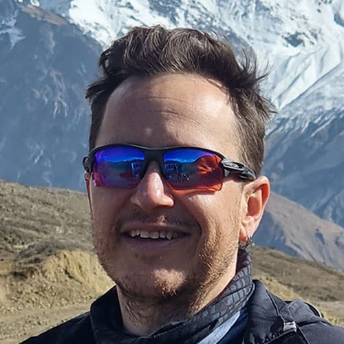 Portrait of Ben A wearing reflective sunglasses, with short curly hair, smiling during our motorcycle tour with snow-capped mountains in the background.
