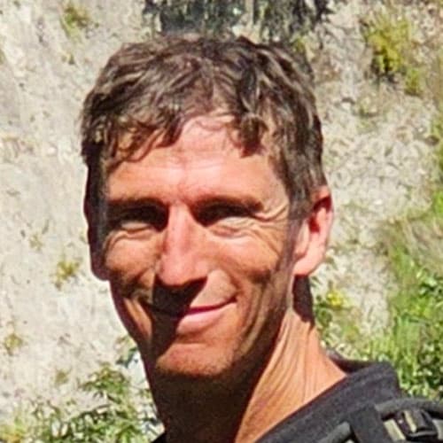 Portrait of Chris B with short brown hair, wearing a backpack strap, smiling against a rocky hillside during our motorcycle tour.