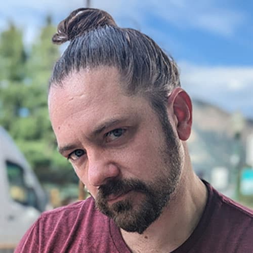 Portrait of Dustin W with a top knot and beard, wearing a maroon shirt, posing during our motorcycle tour with mountains and a vehicle in the background.