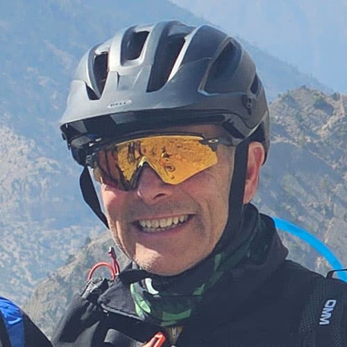 Portrait of Hugh S wearing a black helmet and reflective goggles, smiling with mountain scenery in the background during our motorcycle tour.