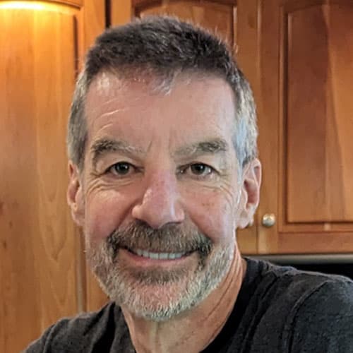 Portrait of Jean-Paul M with short gray hair and a beard, smiling in front of a wooden cabinet.