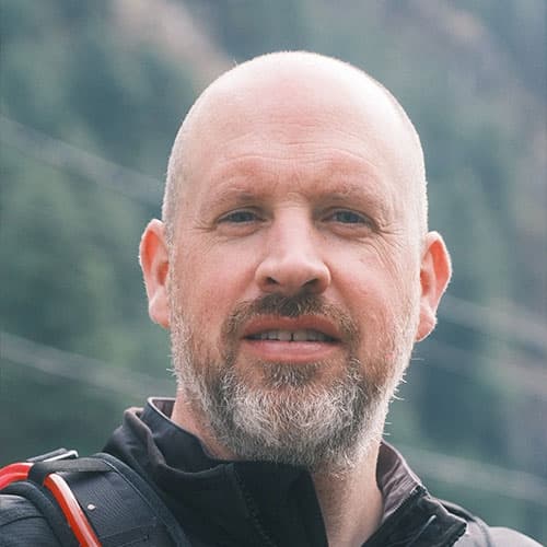 Portrait of Jon H with a shaved head and beard, wearing an outdoor jacket with a backpack strap, in front of a blurred mountain background during our motorcycle tour.