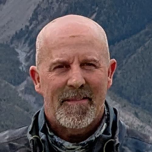 Portrait of Tim F, a man with a bald head and goatee, wearing a neck gaiter, with mountains in the background.